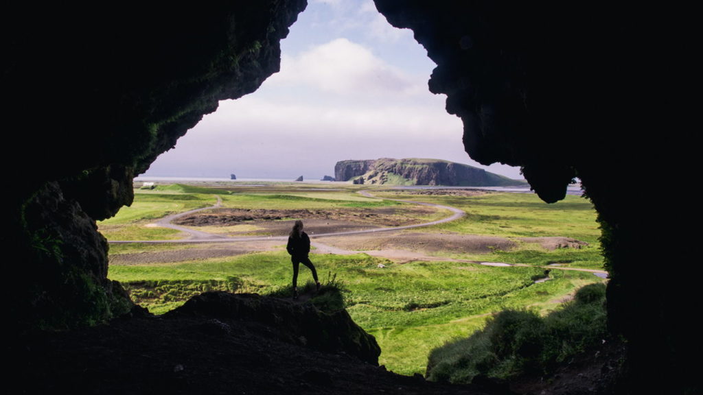 person looking over field from the mouth of a cave